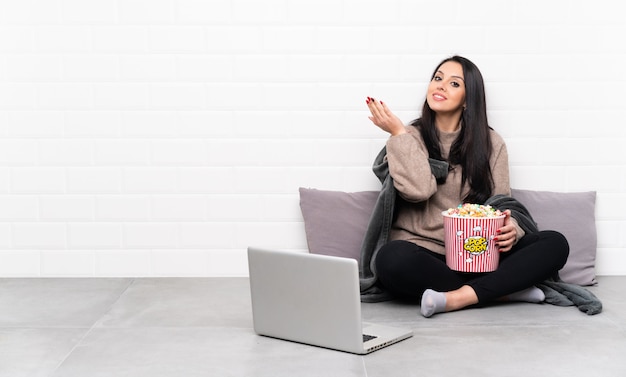 Jonge Colombiaanse vrouw die een kom popcorns vasthoudt en een film laat zien in een laptop die haar handen naar de zijkant uitstrekt om uit te nodigen om te komen