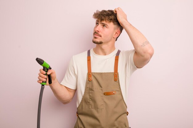 Jonge coole man die zich verbaasd en verward voelt, krabt aan het hoofd van tuinman en slangconcept