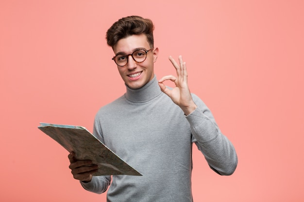 Jonge coole man met een kaart verrast wijzend op zichzelf, breed glimlachend.