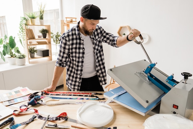 Jonge creatieve man die een decor op een van de werkstukken van een huisdierhalsband drukt of plakt tijdens het gebruik van technische apparatuur