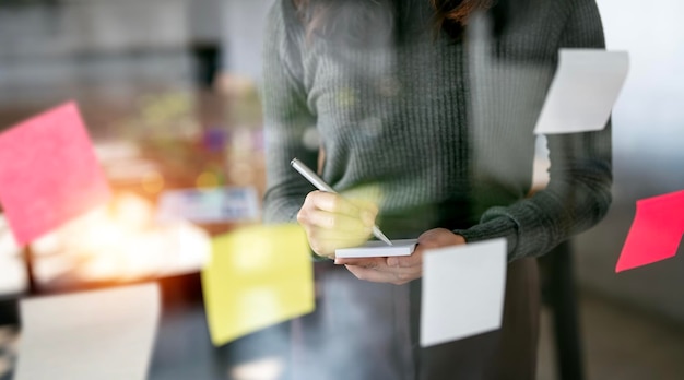 Jonge creatieve zakenvrouw die notitie schrijft en post-it-notities in glazen wand gebruikt om strategiezaken en ontwikkeling tot succes te plannen