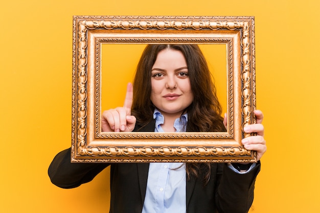 Jonge curvy plus grootte bedrijfsvrouw die een kader houdt dat nummer één met vinger toont.