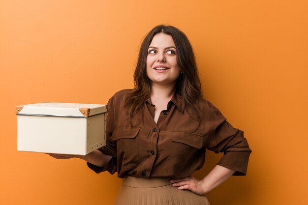 Jonge curvy plus groottevrouw die doos glimlachen houden zeker met gekruiste wapens.