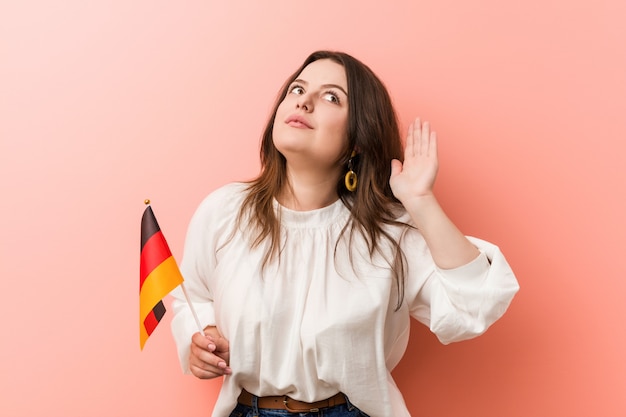 Jonge curvy plus groottevrouw die een vlag houden die van Duitsland aan het luisteren van een roddel probeert.