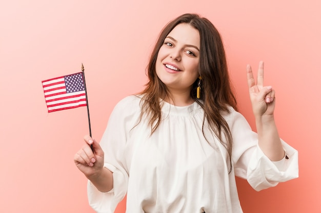 Jonge curvy plus groottevrouw die een vlag houden die van Verenigde Staten overwinningsteken tonen en breed glimlachen.