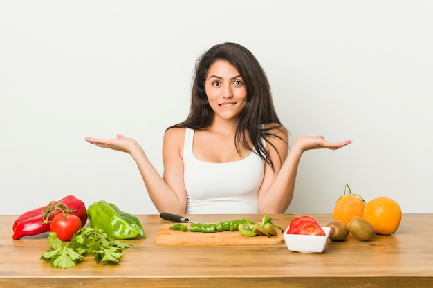 Jonge curvy vrouw die een gezonde maaltijd voorbereidt verwarde en twijfelachtige schouders ophaalt om een exemplaarruimte te houden.