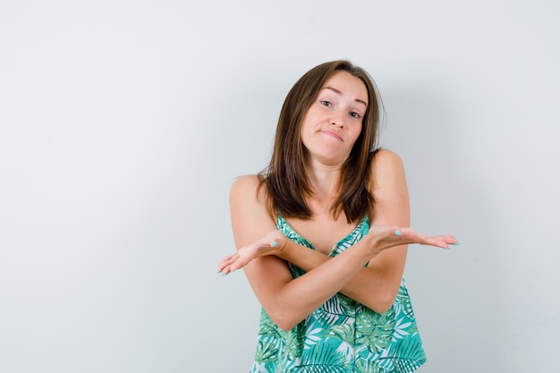 Jonge dame die beide kanten met gekruiste handen in blouse toont en aarzelend kijkt, vooraanzicht.