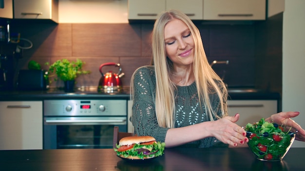 Jonge dame die hamburger verkiest boven salade Aantrekkelijke jonge vrouw die ervoor kiest gezonde hamburger te eten voor...
