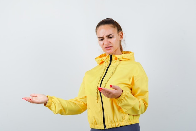 Jonge dame in gele jas die naar haar handpalmen opent en er ontevreden uitziet, vooraanzicht.