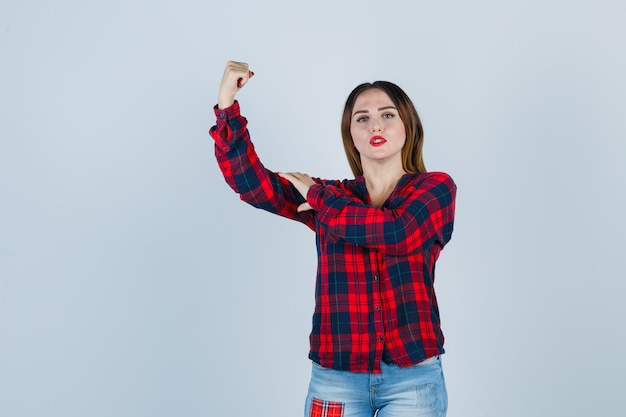 Jonge dame in geruit overhemd, jeans die haar spieren laat zien en er trots uitziet, vooraanzicht.