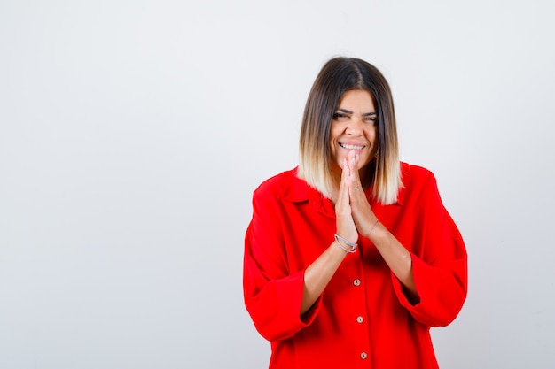 Jonge dame in rood oversized shirt biddend met handen samen om vergeving te vragen en er gelukkig uit te zien, vooraanzicht.