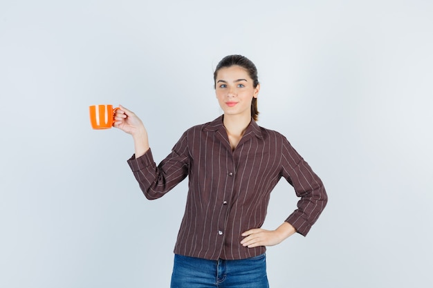 Jonge dame in shirt, spijkerbroek met kop, met hand op taille en schattig, vooraanzicht.