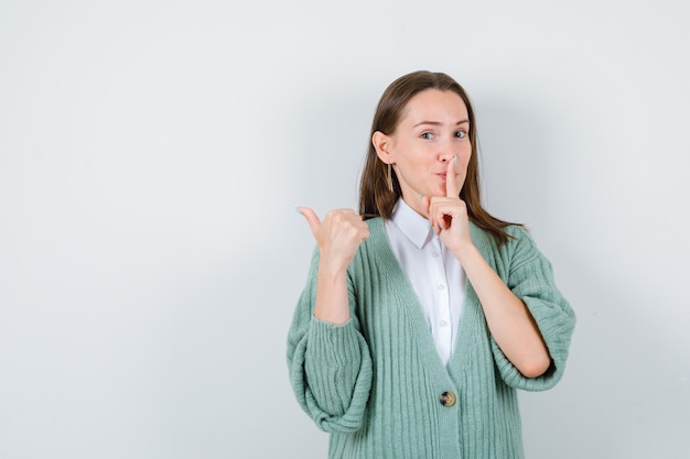 Jonge dame in shirt, vest met stiltegebaar terwijl ze opzij wijst met duim en tevreden kijkt, vooraanzicht.