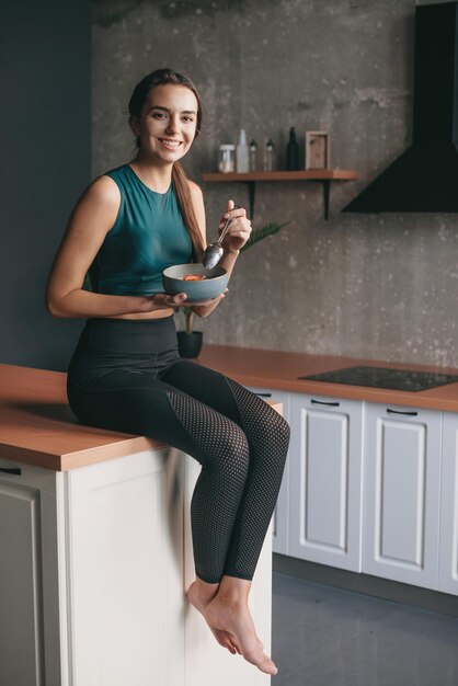 Jonge dame in sportkleding zittend op de keukentafel met een kom cornflakes en met een lepel...