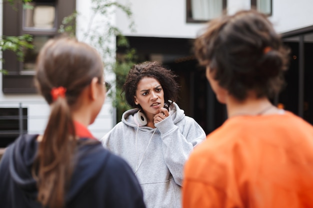 Jonge dame met donker krullend haar in koptelefoon staan en bedachtzaam kijken naar haar vriend terwijl ze tijd doorbrengt met studenten op de binnenplaats van de universiteit