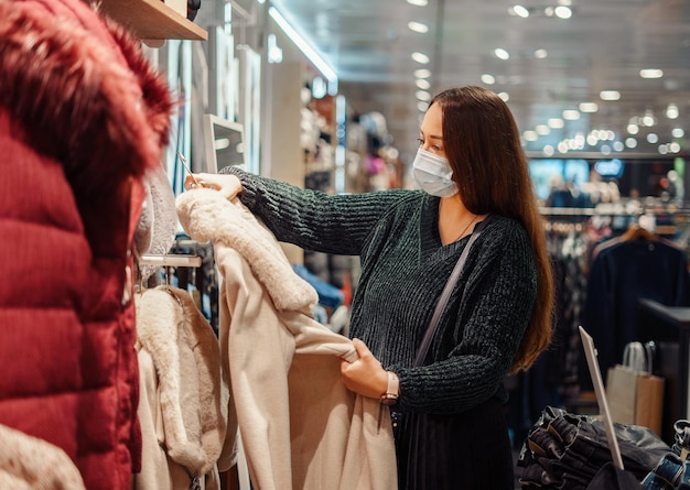 Jonge dame met een wit gezichtsmasker die tijdens een pandemie de warme winterjas in het winkelcentrum controleert