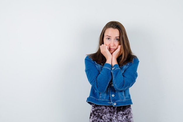 Jonge dame steunt kin op handen in blouse, spijkerjasje en ziet er mooi uit. vooraanzicht.