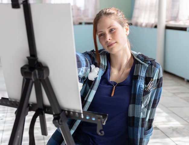 Jonge de kunstenaarsverven van de tienervrouw met olieverven die op de marmeren vloer zitten. Wit canvas en ezel staan op de vloer van marmeren tegels in de kamer met turquoise en lichtgroene muren.