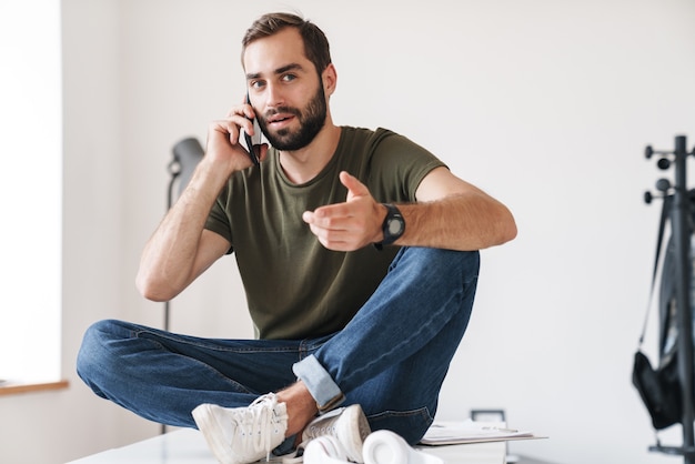 jonge denkende man die op mobiele telefoon praat terwijl hij op een bureau zit met gekruiste benen in een helder kantoor