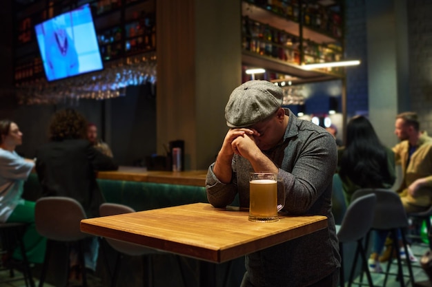 Jonge depressieve man met zijn handen bij zijn gezicht buigend over tafel met een beker bier