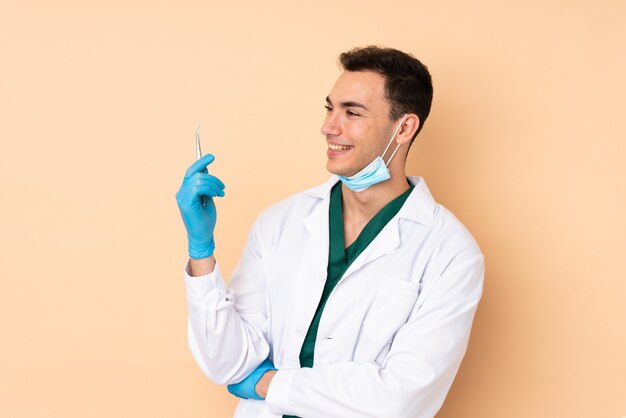 Jonge die de holdingshulpmiddelen van de tandartsmens op beige gelukkig en muur worden geïsoleerd