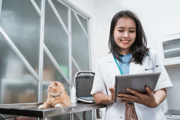 Jonge dierenarts glimlachen tijdens het gebruik van digitale tablet in de buurt van een kat zit op de tafel bij de dierenartskliniek