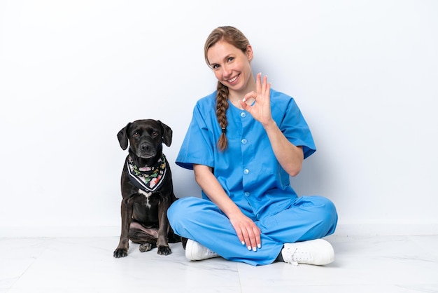 Jonge dierenarts vrouw met hond zittend op de vloer geïsoleerd op een witte achtergrond met ok teken met vingers