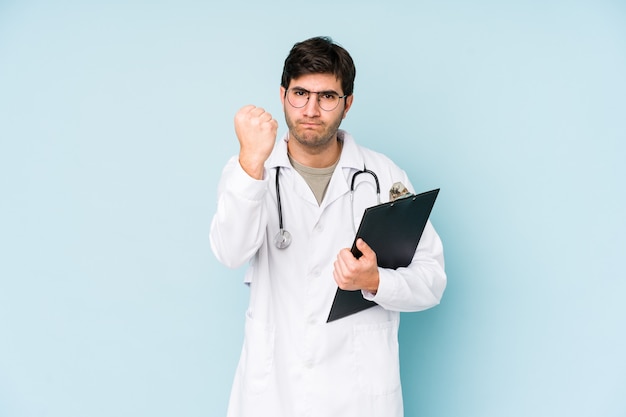 Jonge dokter man geïsoleerd op blauw met vuist naar camera, agressieve gezichtsuitdrukking.