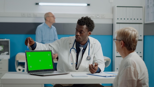 Jonge dokter met laptop met groene schermtechnologie op bureau terwijl hij praat met oudere patiënt in de gezondheidskliniek. Horizontaal apparaat met geïsoleerde mockup-sjabloon en chroma key-achtergrond