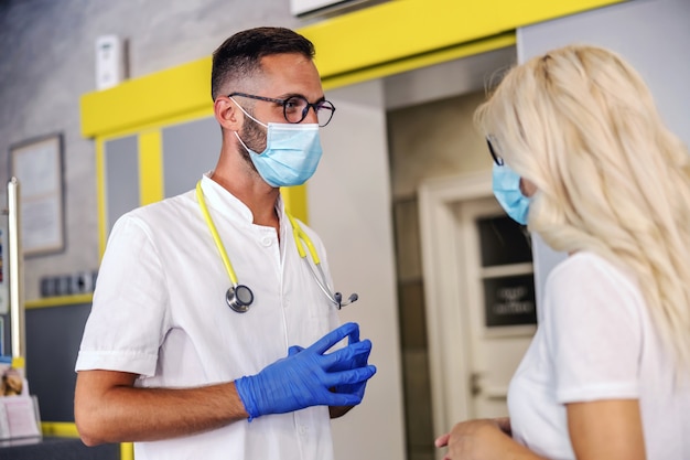 Jonge dokter met rubberen handschoenen, gezichtsmasker in steriel uniform staat in het ziekenhuis en neemt naar een patiënt over de testresultaten van het coronavirus. O