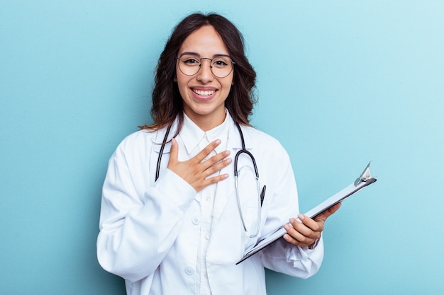 Jonge dokter Mexicaanse vrouw geïsoleerd op blauwe achtergrond lacht hardop met de hand op de borst.