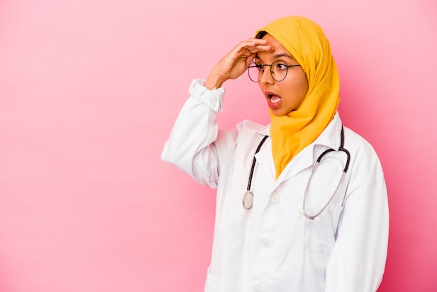 Jonge dokter moslimvrouw geïsoleerd op een roze achtergrond die ver weg kijkt en hand op het voorhoofd houdt.