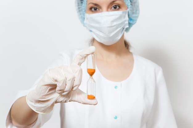 Jonge dokter vrouw met gezichtsmasker, steriele hoed, handschoenen geïsoleerd op een witte achtergrond. Vrouwelijke chirurg arts in medische jurk houdt zandloper. Zorgpersoneel, geneeskundeconcept. De tijd raakt op.