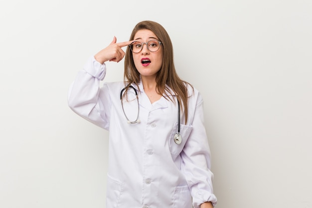 Jonge dokter vrouw tegen een witte muur met een gebaar van teleurstelling met wijsvinger.