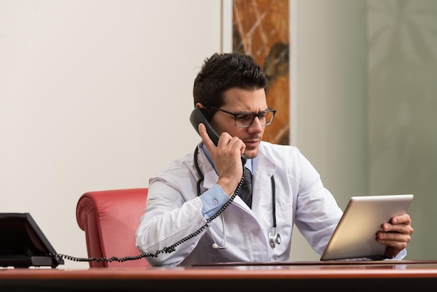 Jonge dokter werkt op zijn computer terwijl hij aan de telefoon is