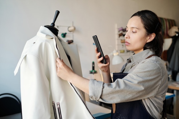 Jonge donkerbruine kleermaker die in werkkleding foto van jasje op smartphone neemt