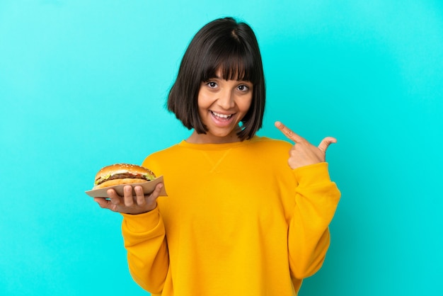 Jonge donkerbruine vrouw die een hamburger over geïsoleerde achtergrond houdt die een duim omhoog gebaar geeft