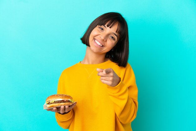 Jonge donkerbruine vrouw die een hamburger over geïsoleerde achtergrond houdt die met gelukkige uitdrukking naar voren wijst