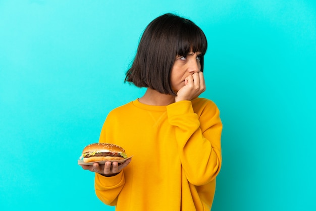 Jonge donkerbruine vrouw die een hamburger over geïsoleerde achtergrond houdt die twijfels heeft