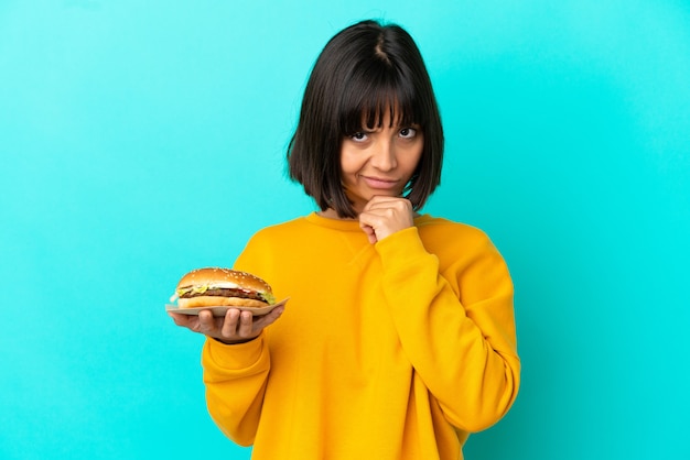 Jonge donkerbruine vrouw die een hamburger over het geïsoleerde denken als achtergrond houdt