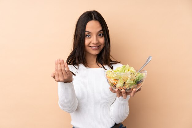 Jonge donkerbruine vrouw die een salade houdt uitnodigend om met hand te komen. Blij dat je bent gekomen