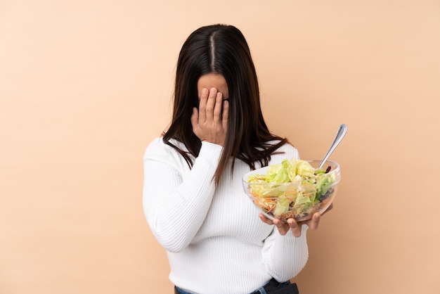 Jonge donkerbruine vrouw die een salade over geïsoleerde muur met vermoeide en zieke uitdrukking houdt