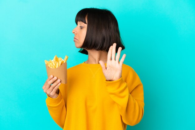 Jonge donkerbruine vrouw die gefrituurde chips over een geïsoleerd blauw oppervlak houdt en een stopgebaar maakt en teleurgesteld is?