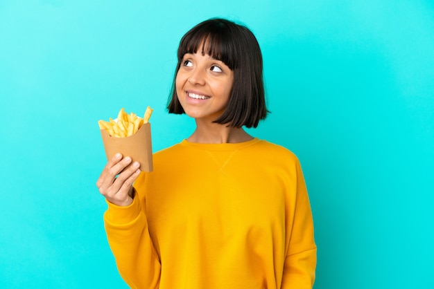 Jonge donkerbruine vrouw die gefrituurde chips over een geïsoleerde blauwe achtergrond houdt en een idee denkt terwijl ze omhoog kijkt