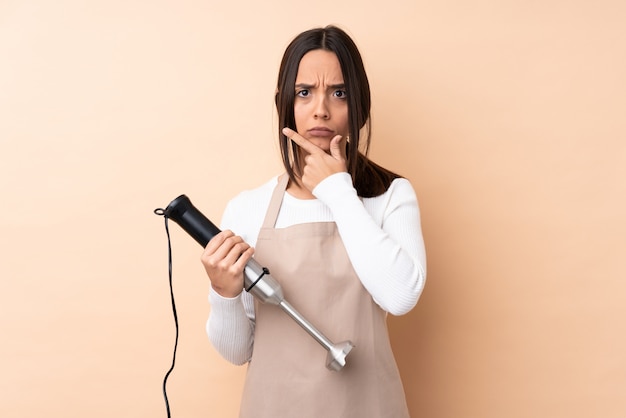 Jonge donkerbruine vrouw die handmixer over het geïsoleerde muur denken gebruikt