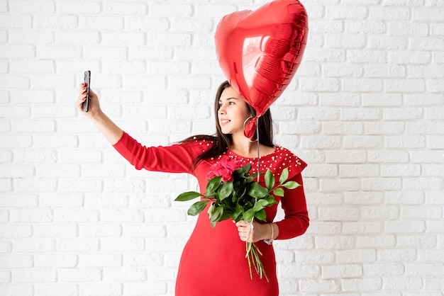 Jonge donkerbruine vrouw die in rode kleding een rode hartballon en bloemen houdt