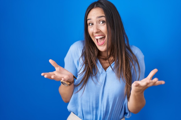 Jonge donkerbruine vrouw die zich over blauwe achtergrond bevindt die gek en verbaasd viert voor succes met opgeheven armen en open ogen schreeuwend opgewonden winnaarconcept