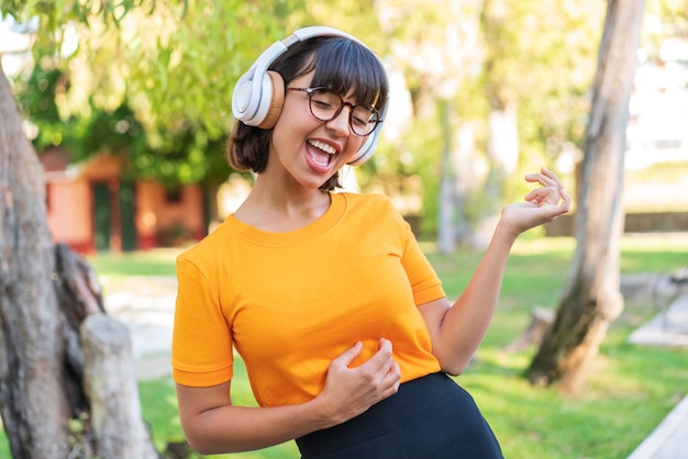 Jonge donkerbruine vrouw in het park die muziek luistert en gitaargebaar doet