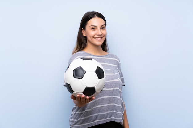 Jonge donkerbruine vrouw over geïsoleerde blauwe muur die een voetbalbal houdt