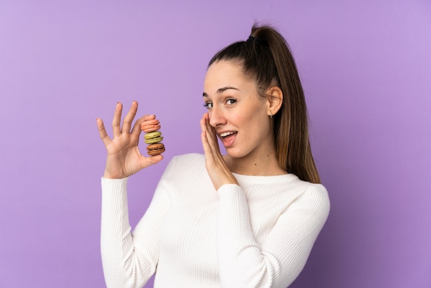 Jonge donkerbruine vrouw over geïsoleerde purpere muur die kleurrijke Franse macarons houden en iets fluisteren